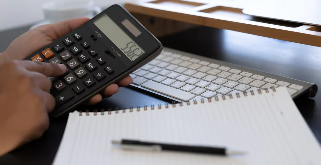 imagem mostra alguém usando uma calculadora, um teclado de computador e um caderno com uma caneta