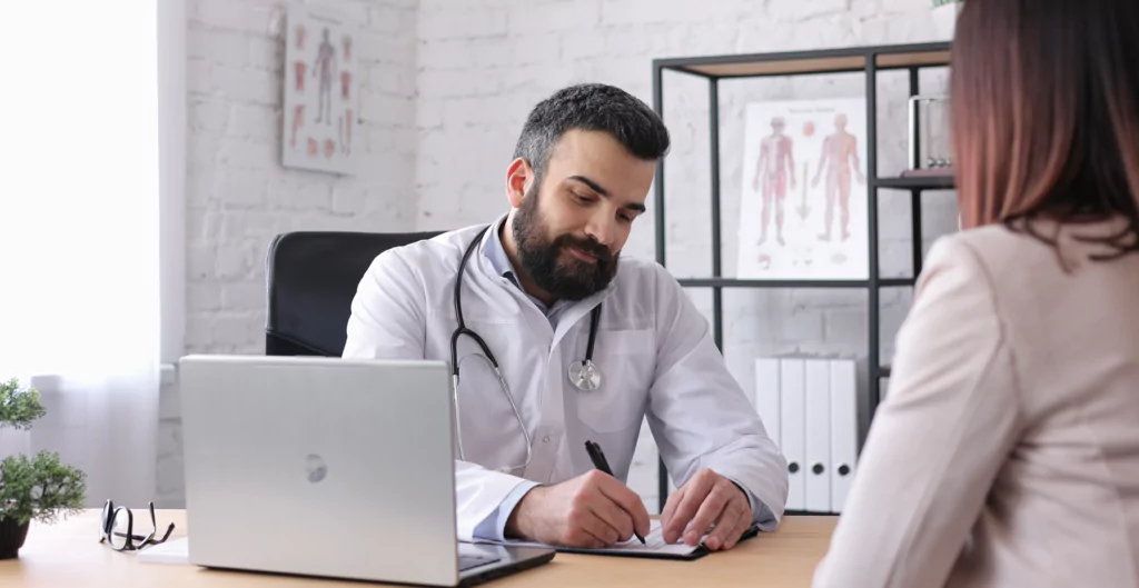 imagem mostra um médico sentado em frente a um notebook e fazendo anotações em um papel. A sua frente, uma mulher está sentada 