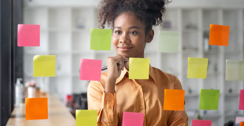 mulher em frente a um vidro cheio de post it com a mão no queixo e expressão de satisfeita 