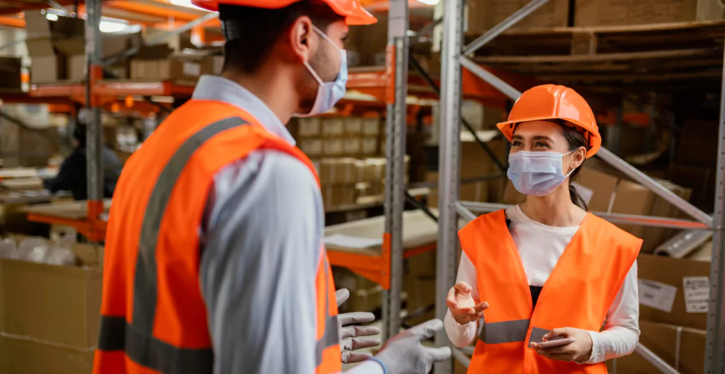 Funcionários de armazém conversando com EPIs e máscaras, praticando comportamento seguro.