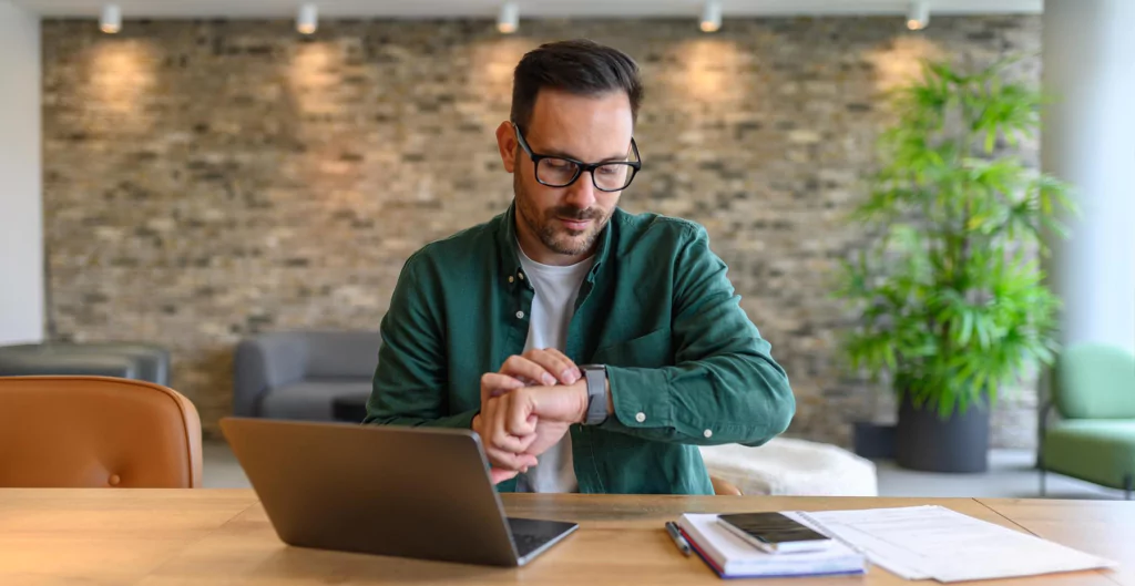 homem em um escritório sentado de frente para um notebook e olhando o seu relógio de pulso 