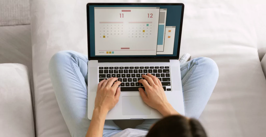 imagem de uma mulher sentada em um sofá com pernas cruzadas e com o calendário do notebook aberto 