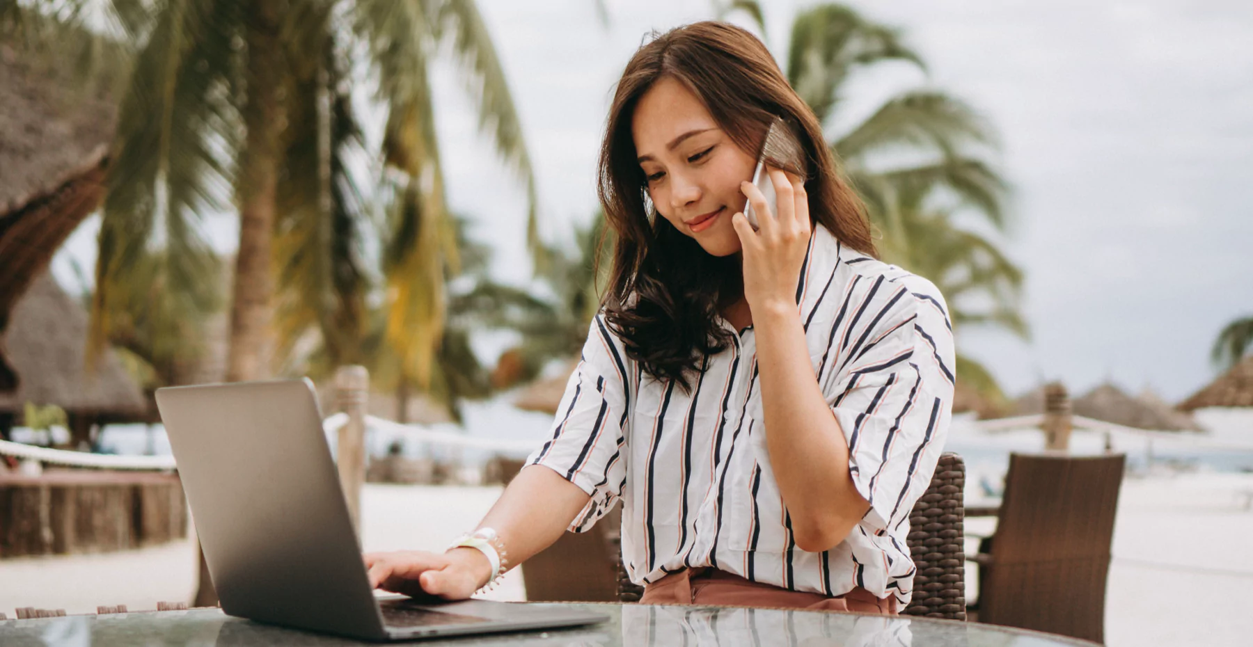 mulher sentada em uma mesa na praia, mexendo no notebook e falando no celular