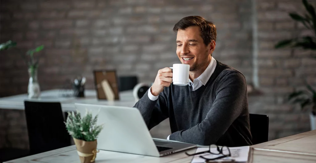 homem sorridente segurando uma xícara de café e sentado em frente a um notebook 