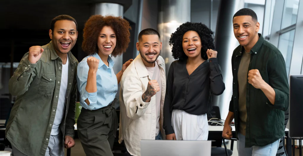 Equipe diversa de colegas vibrando e demonstrando entusiasmo no ambiente de trabalho.