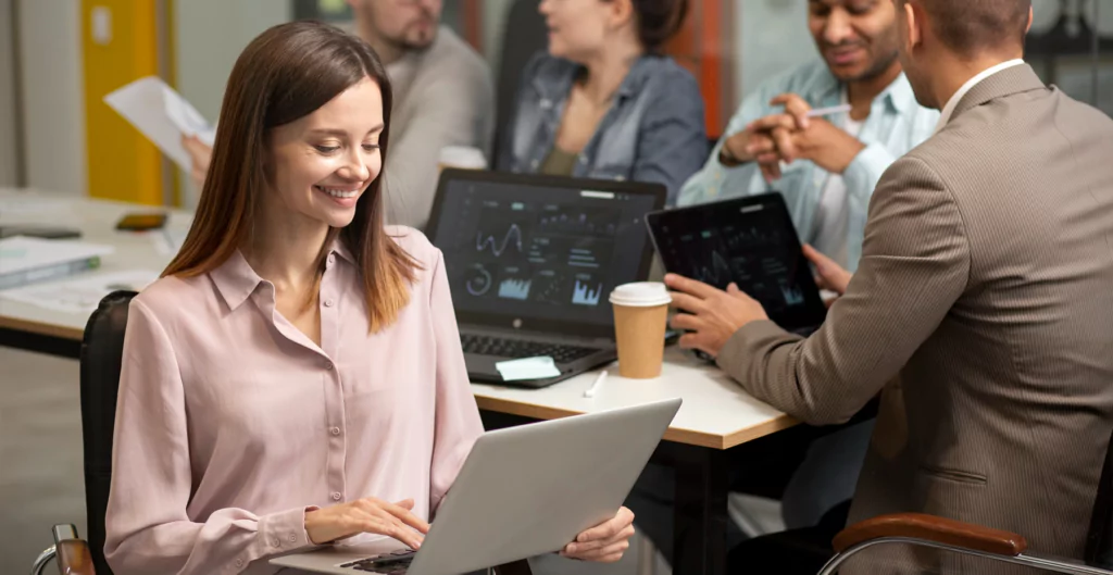 Mulher sorridente trabalhando em laptop, com colegas ao fundo discutindo dados.