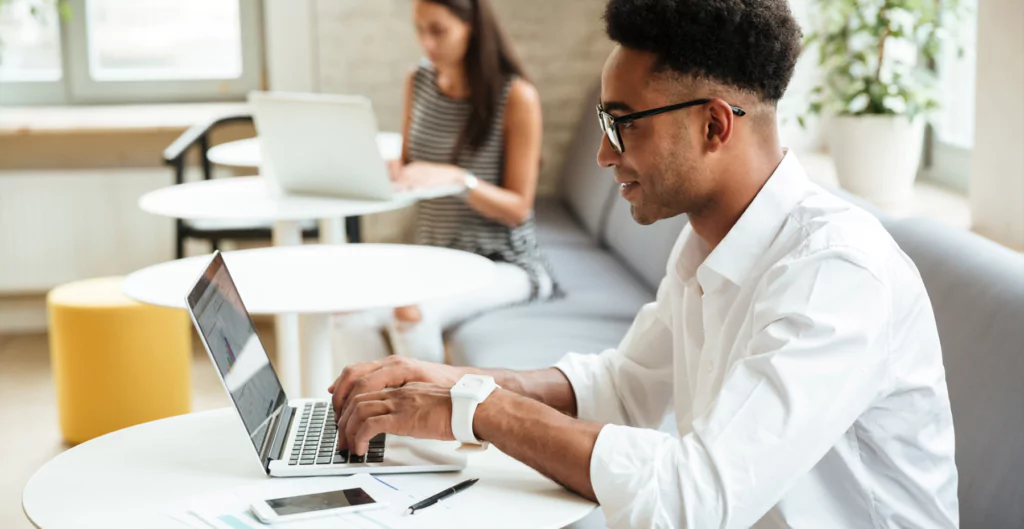 Homem negro sentado em uma pequena mesa redonda com notebook, enquanto trabalha sentado em um sofá 