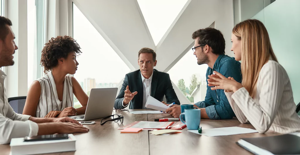 Mesa de negociação empresarial, com 5 pessoas, com canecas, notebooks e documentos 