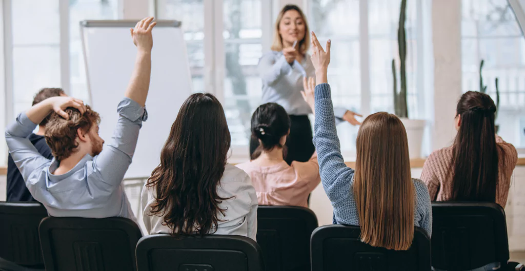 Grupo de pessoas em treinamento corporativo, com participantes levantando as mãos para perguntas, destacando a educação e desenvolvimento no RH ideal.