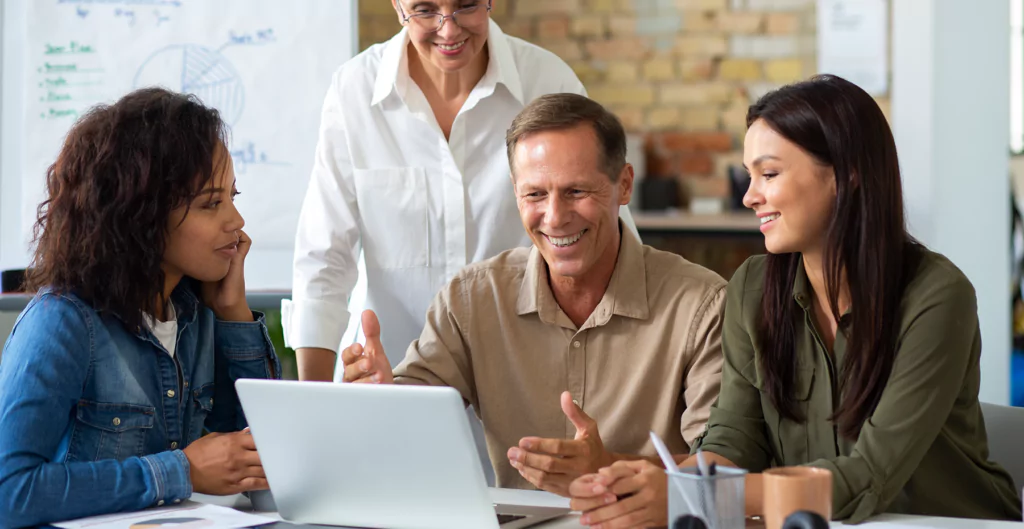 Equipe reunida em torno de um laptop, discutindo e sorrindo, enfatizando trabalho em equipe e ambiente positivo no RH ideal.