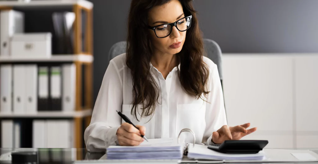 Mulher analisando documentos e usando calculadora em escritório, simbolizando cálculo e organização financeira.