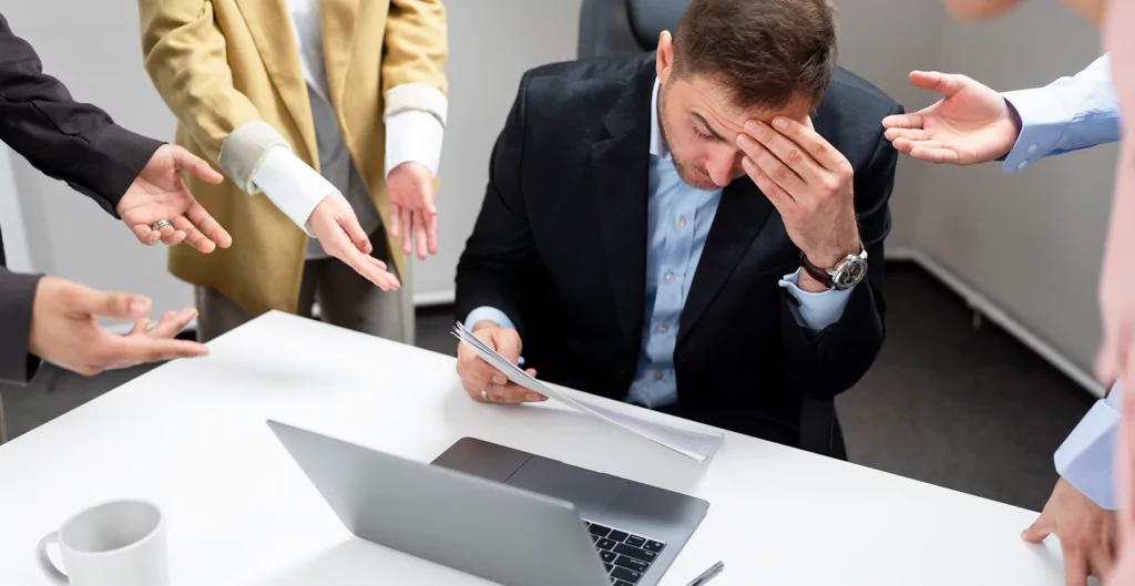 Homem sobrecarregado no trabalho, cercado por colegas exigentes.