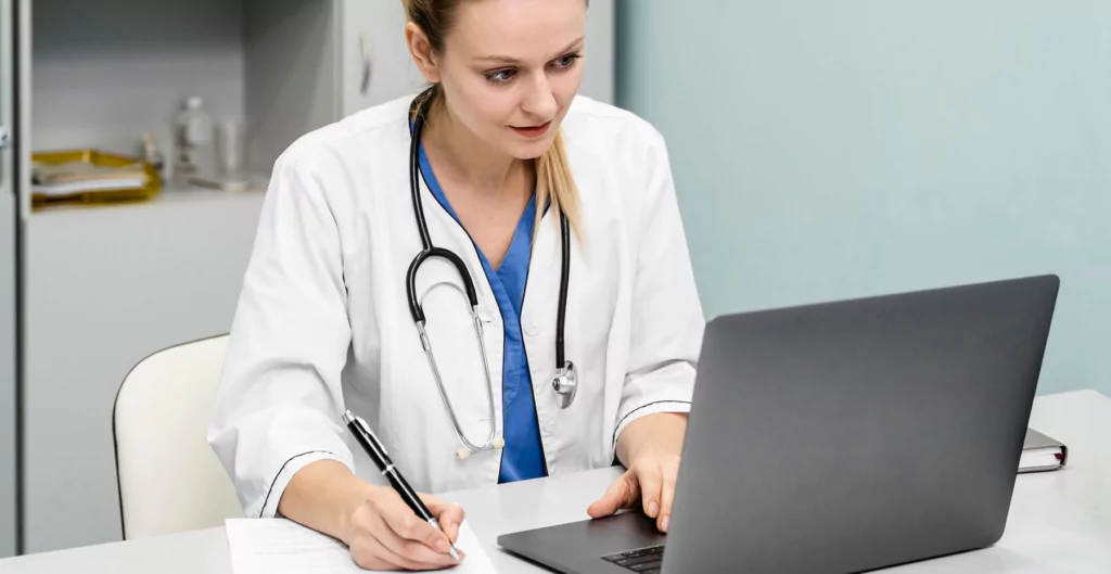 médica sentada com jaleco, digitando no computador e anotando em um papel, representando o atesta CFM