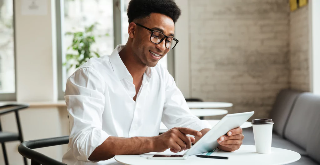 Homem negro, com blusa branca, em sentado com um tablet na mão e sorrindo