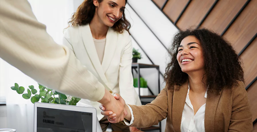 Ambiente corporativo de colaboração e confiança, com duas mulheres profissionais interagindo de forma positiva. Uma delas, sorridente, aperta a mão de uma pessoa fora do quadro, enquanto outra observa amigavelmente ao fundo. 