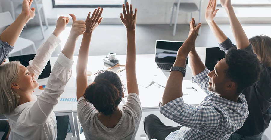 Ambiente corporativo, com cinco pessoas com os braços levantados em sinal de motivação com o trabalho 