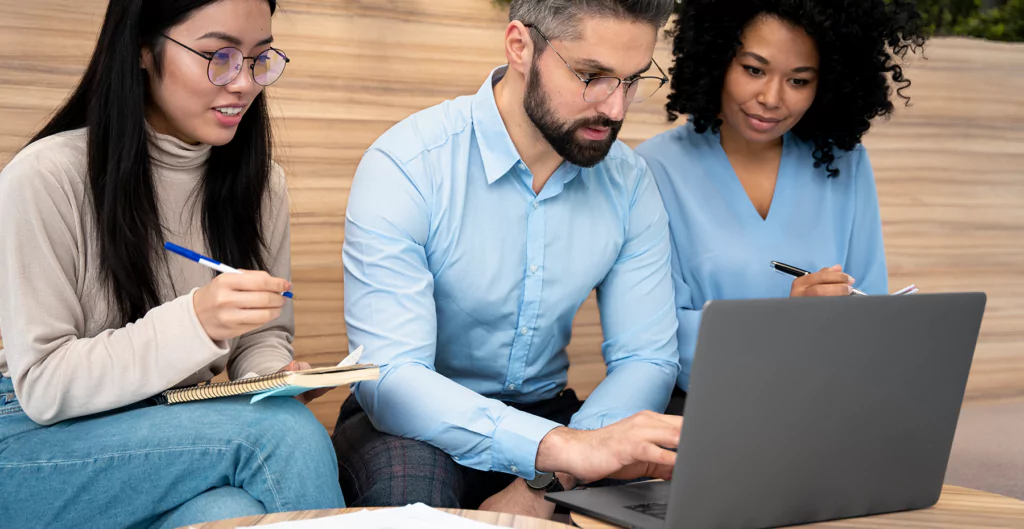 três pessoas sentadas olhando um notebook e fazendo anotações 