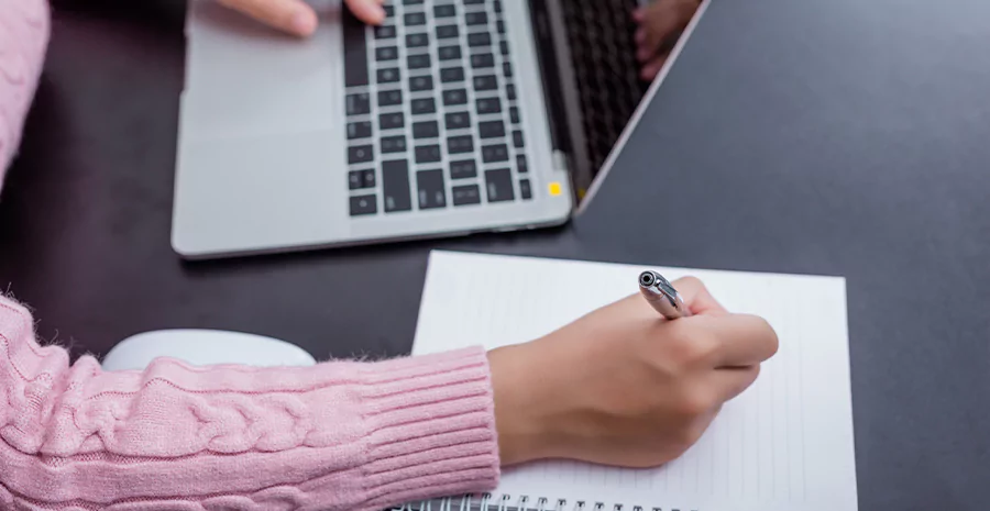 pessoa escrevendo algo em um caderno com o notebook ao lado. Imagem representa checklist de admissão de funcionários