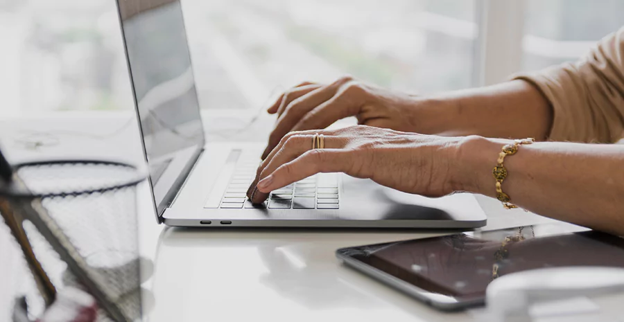 Mãos com acessórios, digitando em um notebook, em uma mesa com canetas e ipad.