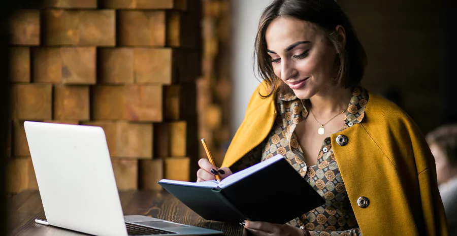 mulher sentada fazendo anotações em um caderno com um notebook na mesa. Imagem representa profissionais multifuncionais