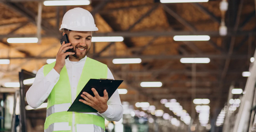 Homem com roupas e equipamentos de segurança de trabalho, com uma fábrica ao fundo