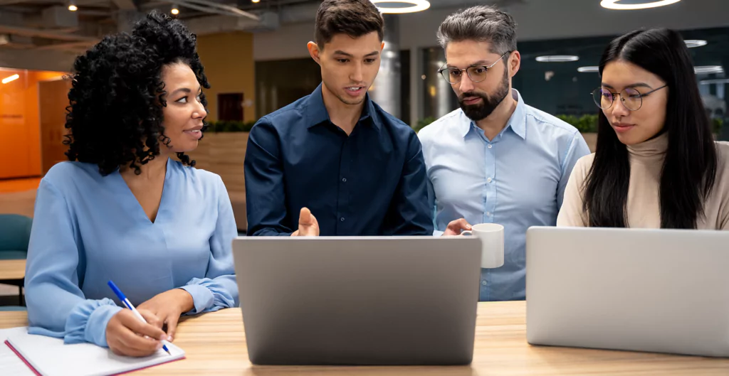 Quatro pessoas analisando arquivos em dois notebooks com ambiente corporativo no fundo