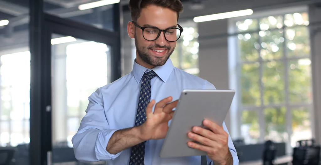 Homem com roupas sociais sorrindo com um tablet na mão, com um escritório corporativo ao fundo 