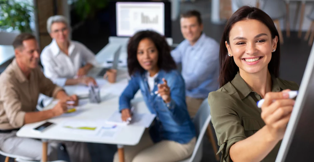 Mulher escrevendo em quadro de ideias com grupo de pessoas ao fundo em uma reunião