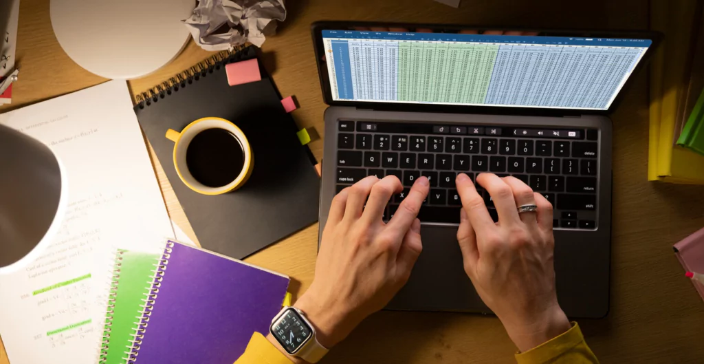 Mãos digitando em notebook com tela em uma longa planilha, cadernos e uma xícara de café ao lado