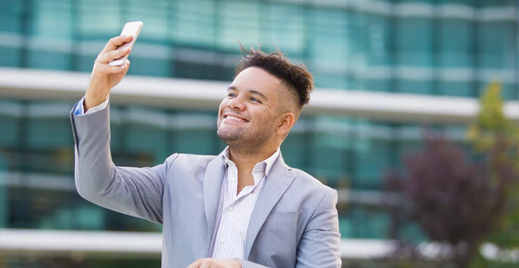 Homem registro ponto com celular através do reconhecimento facial com prédio ao fundo 