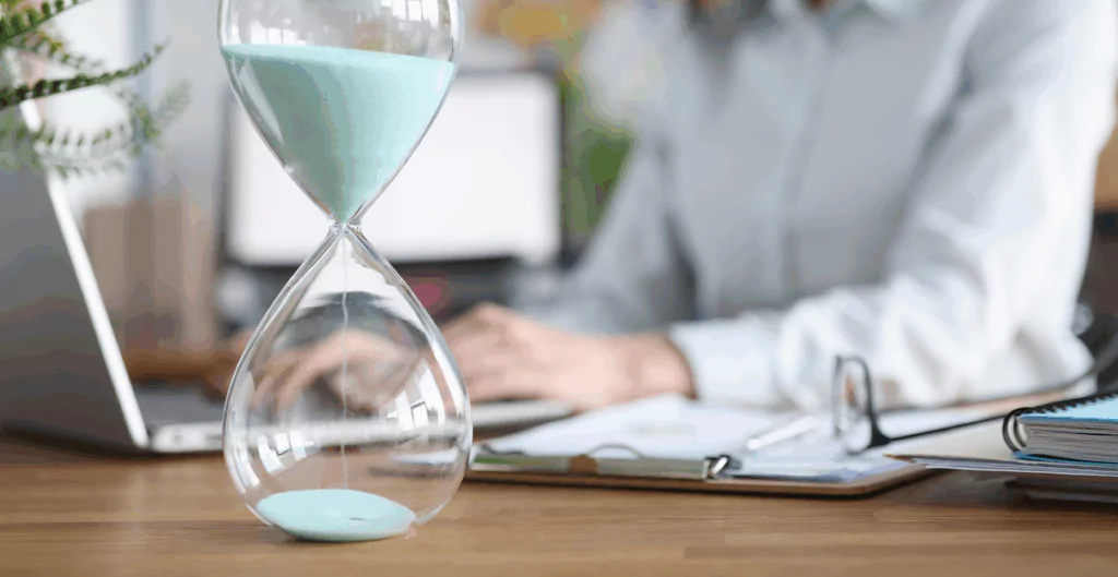 Relógio de areia na mesa de trabalho com uma pessoa ao fundo usando laptop e documentos em ambiente de escritório