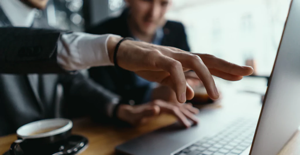Homens de terno colaborando em um escritório moderno, analisando informações em um laptop enquanto tomam café.