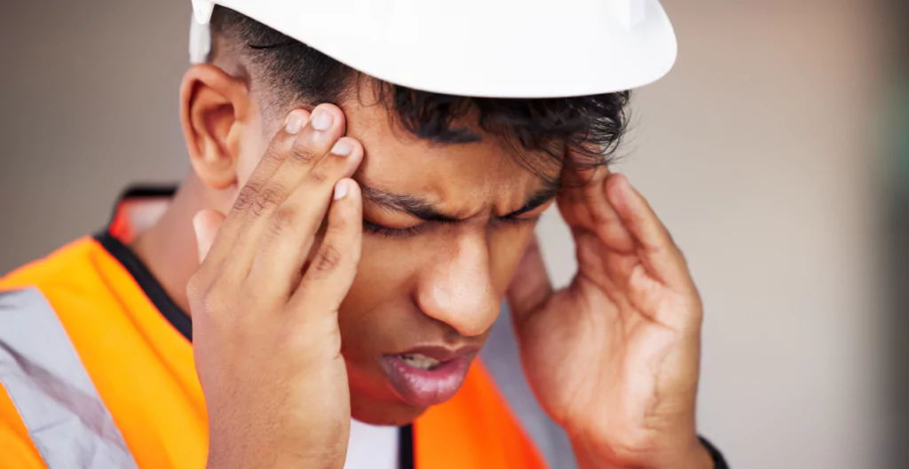 Trabalhador da construção civil com expressão de dor e mãos na cabeça, simbolizando sintomas de doença ocupacional compatível com enquadramento no NTEP.