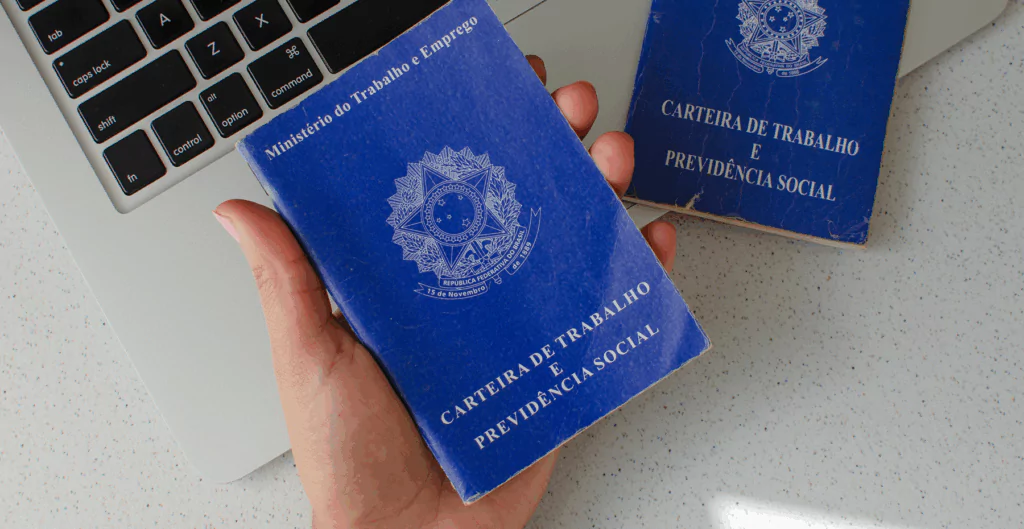 Pessoa segurando uma carteira de trabalho e previdência social azul, símbolos do Brasil visíveis, colocando documentos sobre uma mesa com um laptop ao lado, representando busca por emprego e direitos trabalhistas.