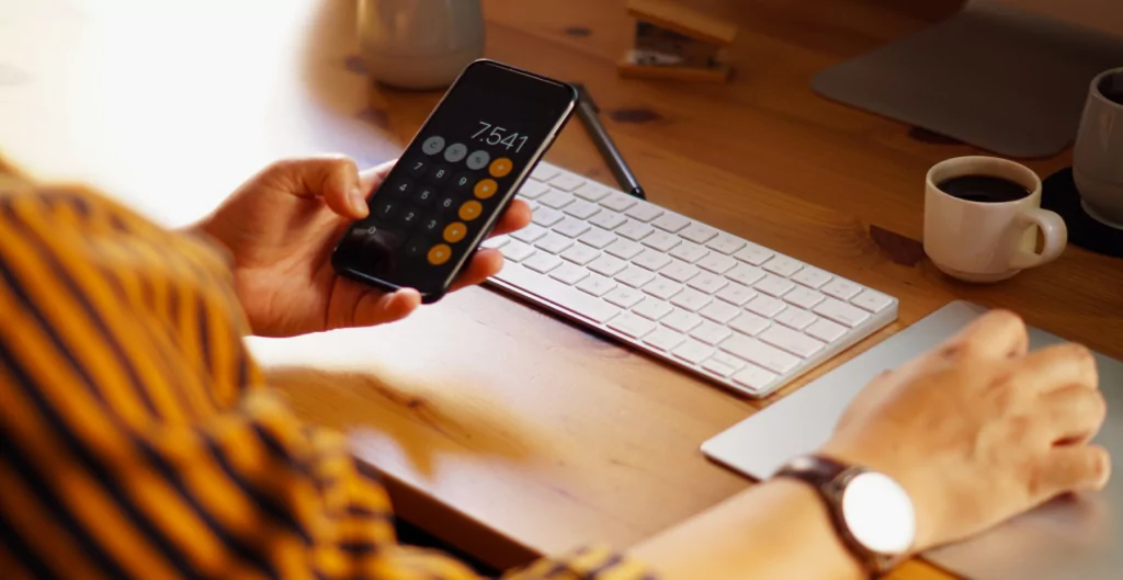 Pessoa utiliza aplicativo de calculadora no celular em mesa de madeira com teclado e café.