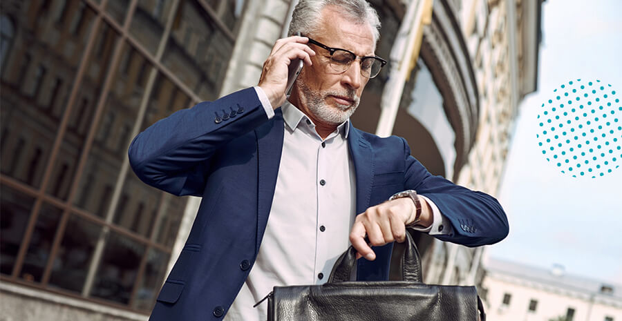 imagem de um homem na frente de um prédio segurando uma maleta, olhando o relógio e falando ao celular preocupado